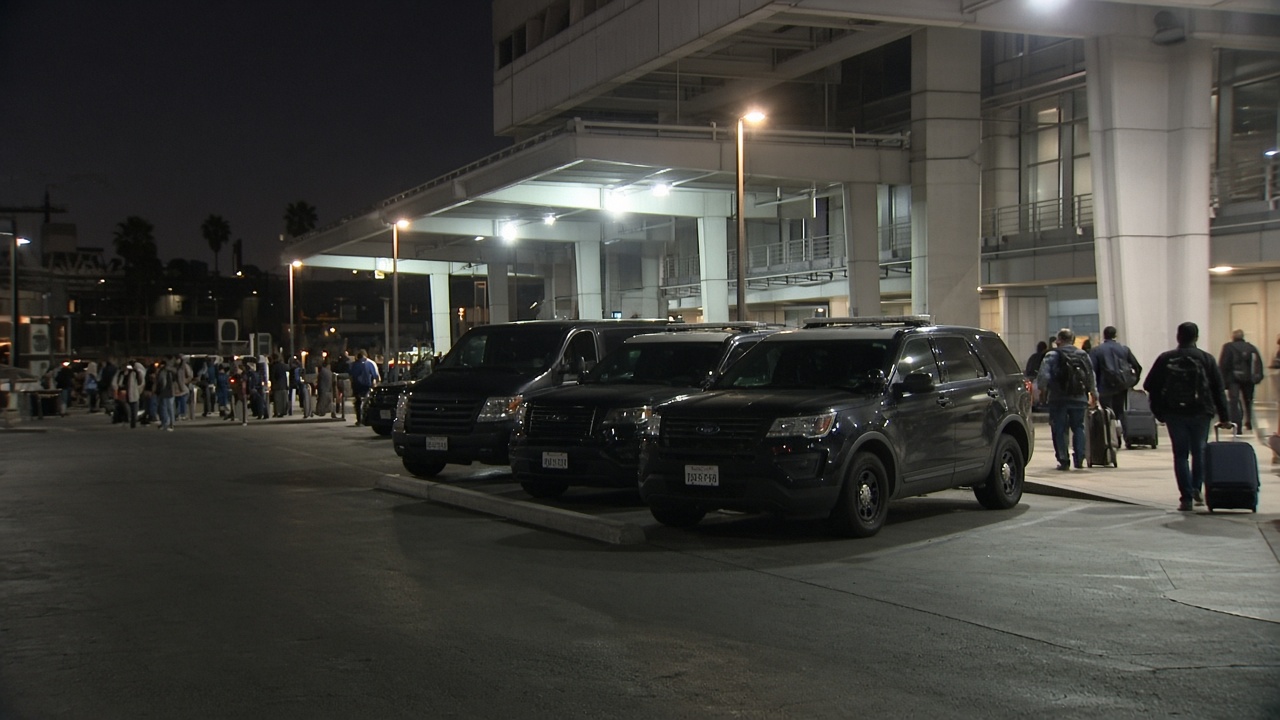 Federal law enforcement vehicles parked near an international terminal at Los Angeles International Airport as travellers wheel luggage under evening floodlights.