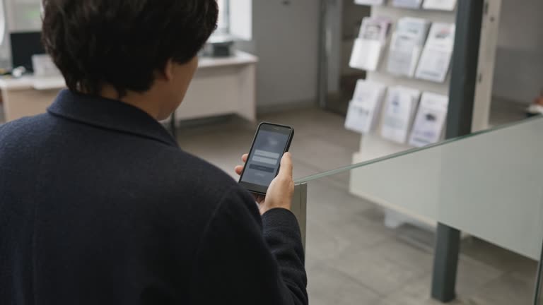 A customer holding a smartphone with a generic banking app login screen at a regional bank lobby, matte floor tiles, brochure rack blurred, no logos or readable text