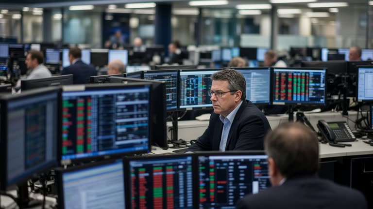 Interior of a London stock exchange dealing room with traders at desks and multiple market screens showing red and green numbers, fluorescent office lighting, shallow depth of field, no readable text or logos