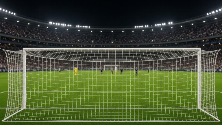 Night football match at a Australian stadium with green pitch, goal net in foreground and blurred crowd lights, no logos or readable text