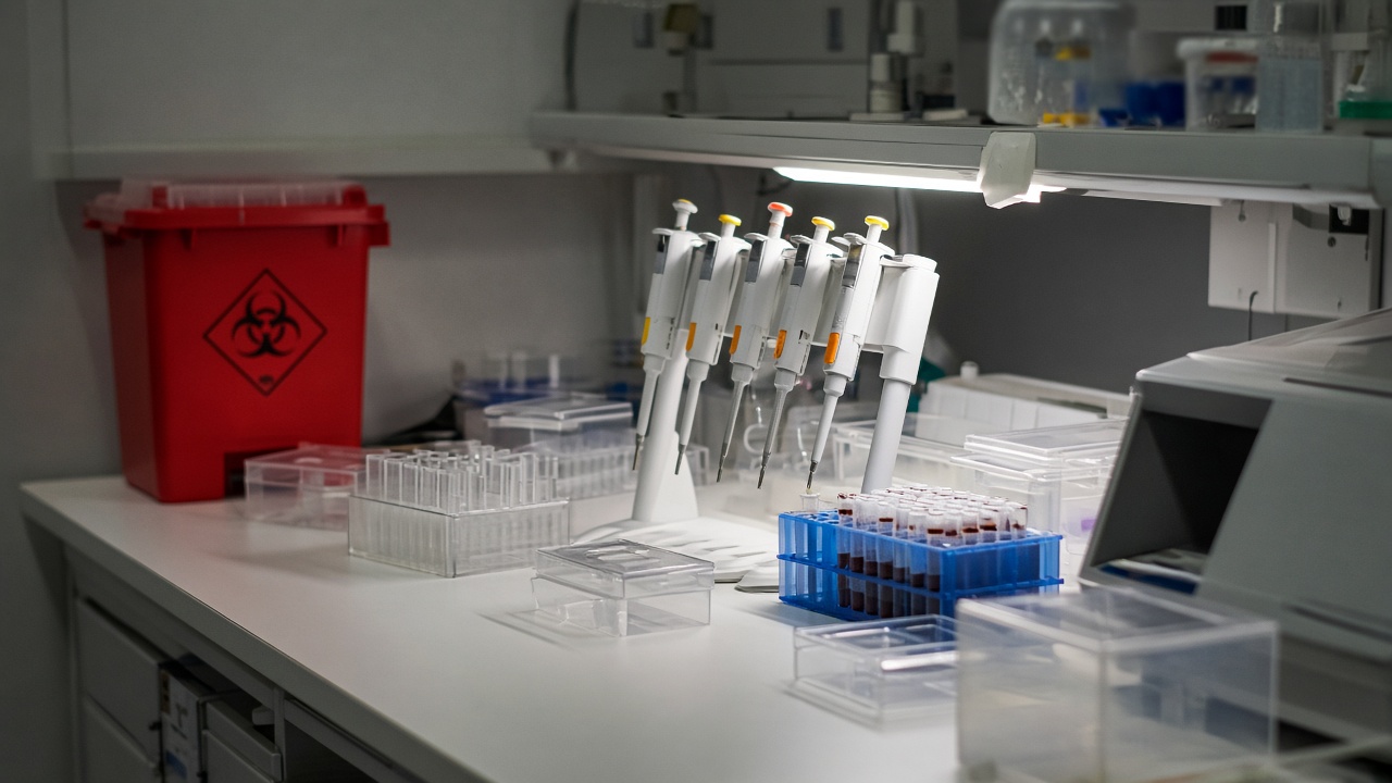 A hospital laboratory bench with pipettes and sample racks under bright task lighting, biohazard bin in the corner, no logos or readable text