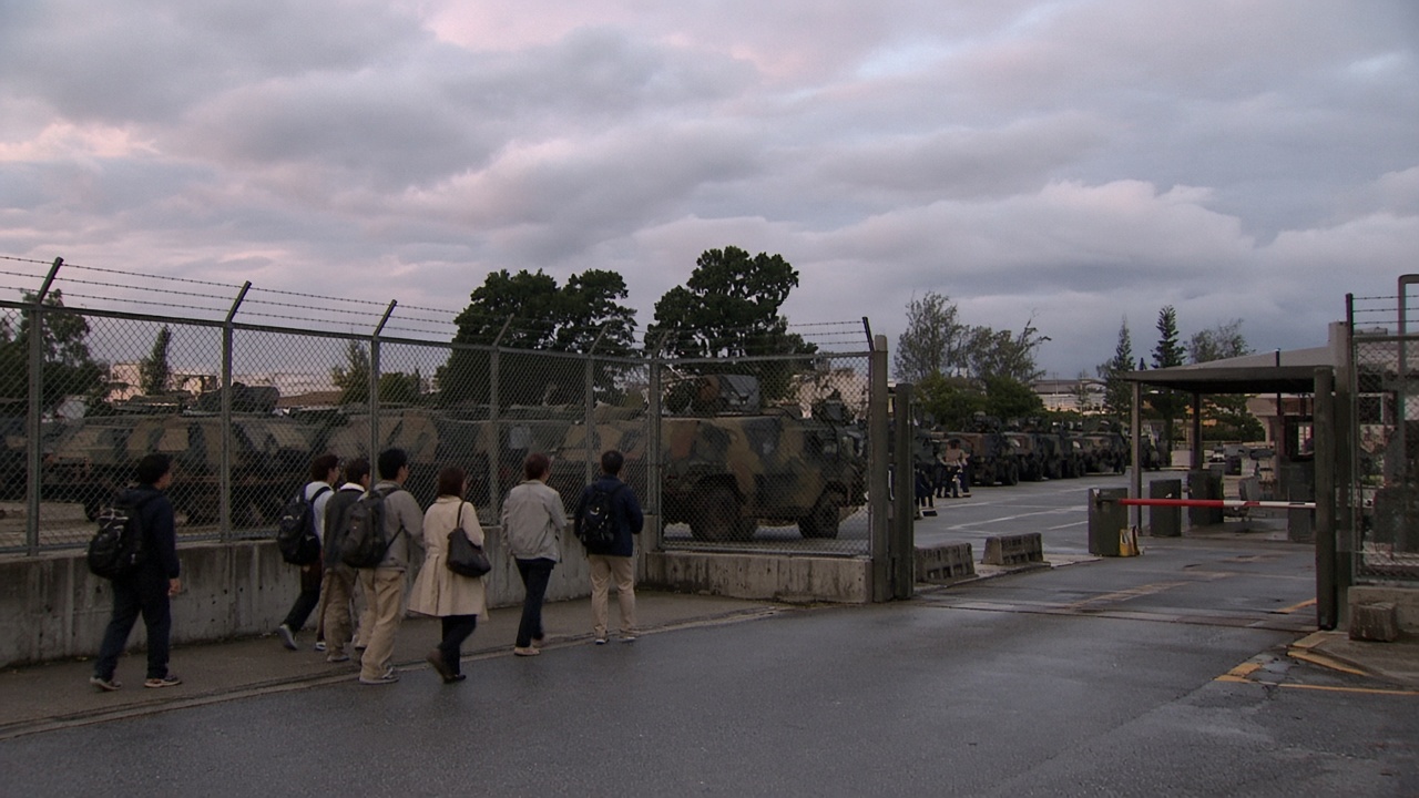 U.S. Marine Corps vehicles parked inside a fenced Okinawa base at dusk as Japanese civilians walk past security gates under overcast skies