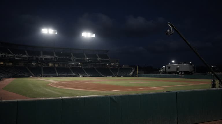 Night exterior of a baseball stadium with floodlights on empty infield, broadcast crane silhouette, production truck parked beyond outfield fence, no logos or readable text