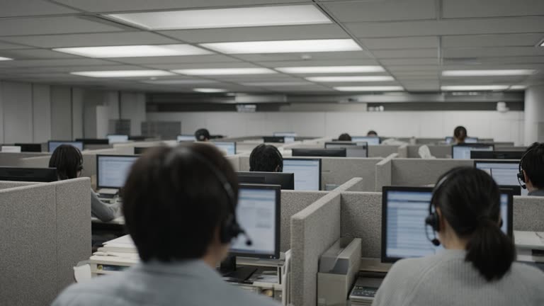 Japanese bank call-center floor with agents wearing headsets viewed from behind, monitors showing blurred text, overhead fluorescent panels, no logos or readable text