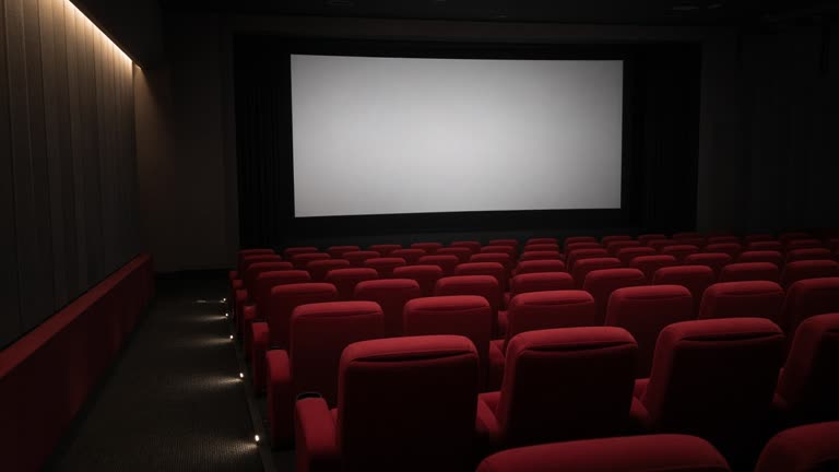 Empty multiplex auditorium with red reclining seats facing a blank screen before the first show, dim aisle lighting, no logos or readable text