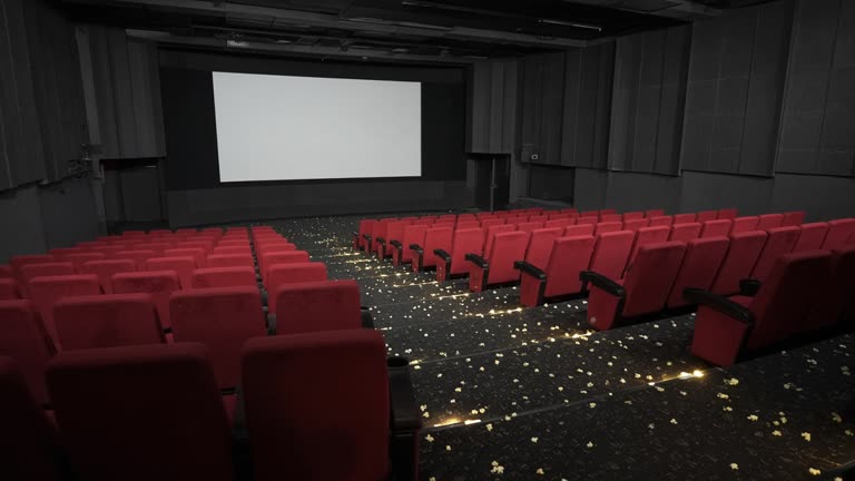 Empty multiplex auditorium with red fabric seats and a blank white screen, dim aisle lighting and scattered popcorn on carpet, no logos or readable text