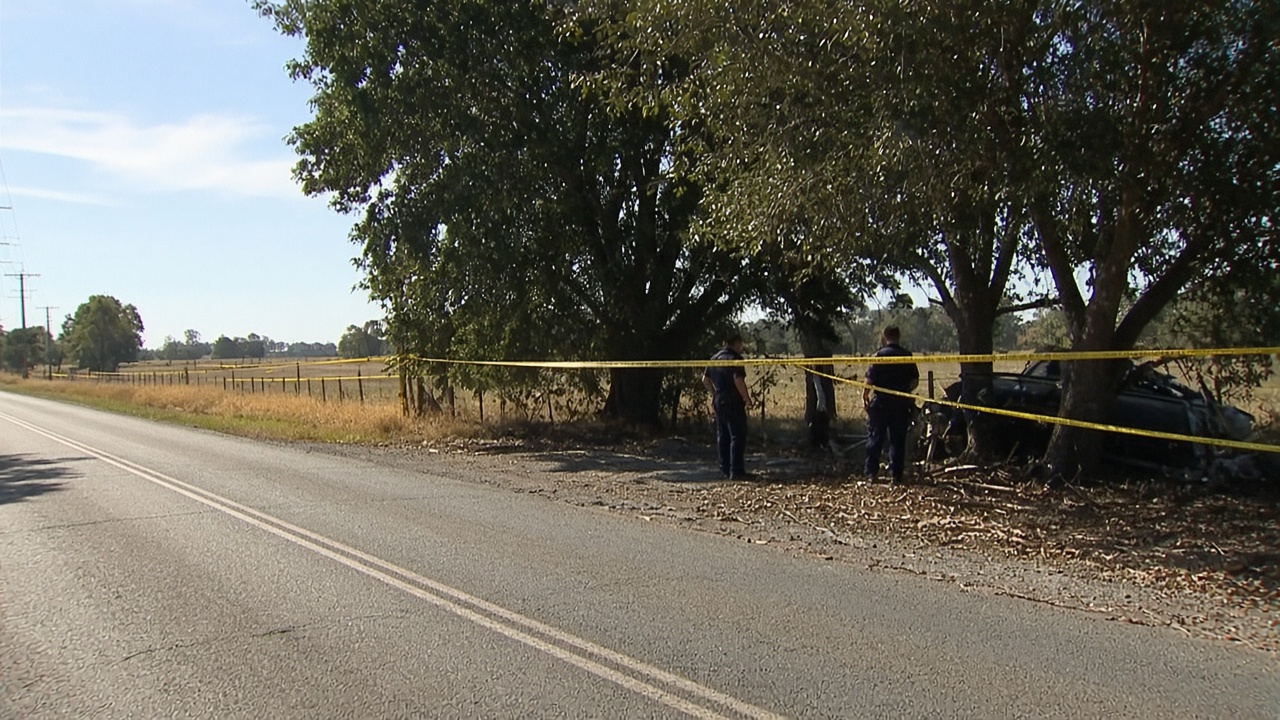 Rural NSW road with yellow police tape fluttering near a tree-lined crash site under harsh afternoon sun, officers silhouetted at distance