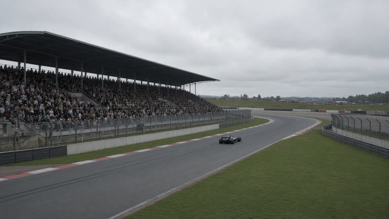 Racing circuit grandstand overlooking a gray asphalt track with a single open-wheel car in the distance, overcast sky, no logos or readable text on car or banners