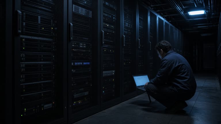 Rows of server racks in a dim data center aisle with a technician on a laptop under cold blue emergency lighting