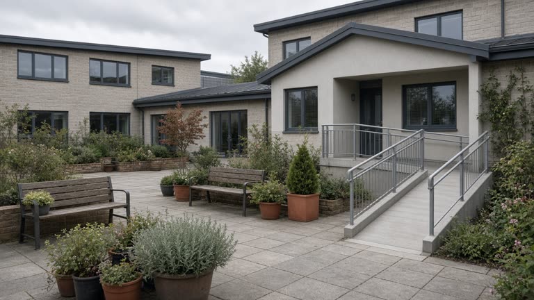 Modern low-rise care home courtyard with benches and potted plants, wheelchair ramp, overcast daylight, no people or readable signage