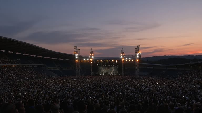 Outdoor stadium concert stage with truss lighting towers seen from the rear seating bowl at dusk, distant crowd as soft silhouettes, no logos or readable text